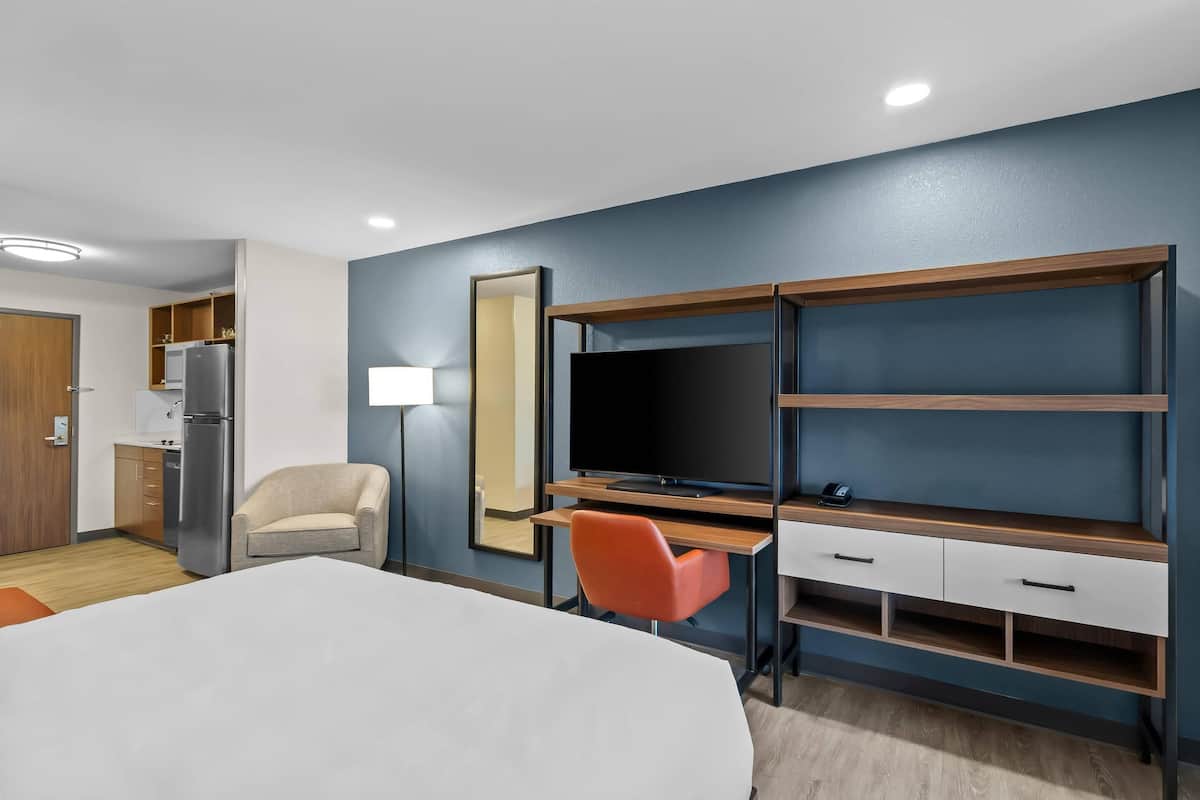 A hotel room with a bed, a desk with a chair, a TV, a mirror, and a kitchenette.