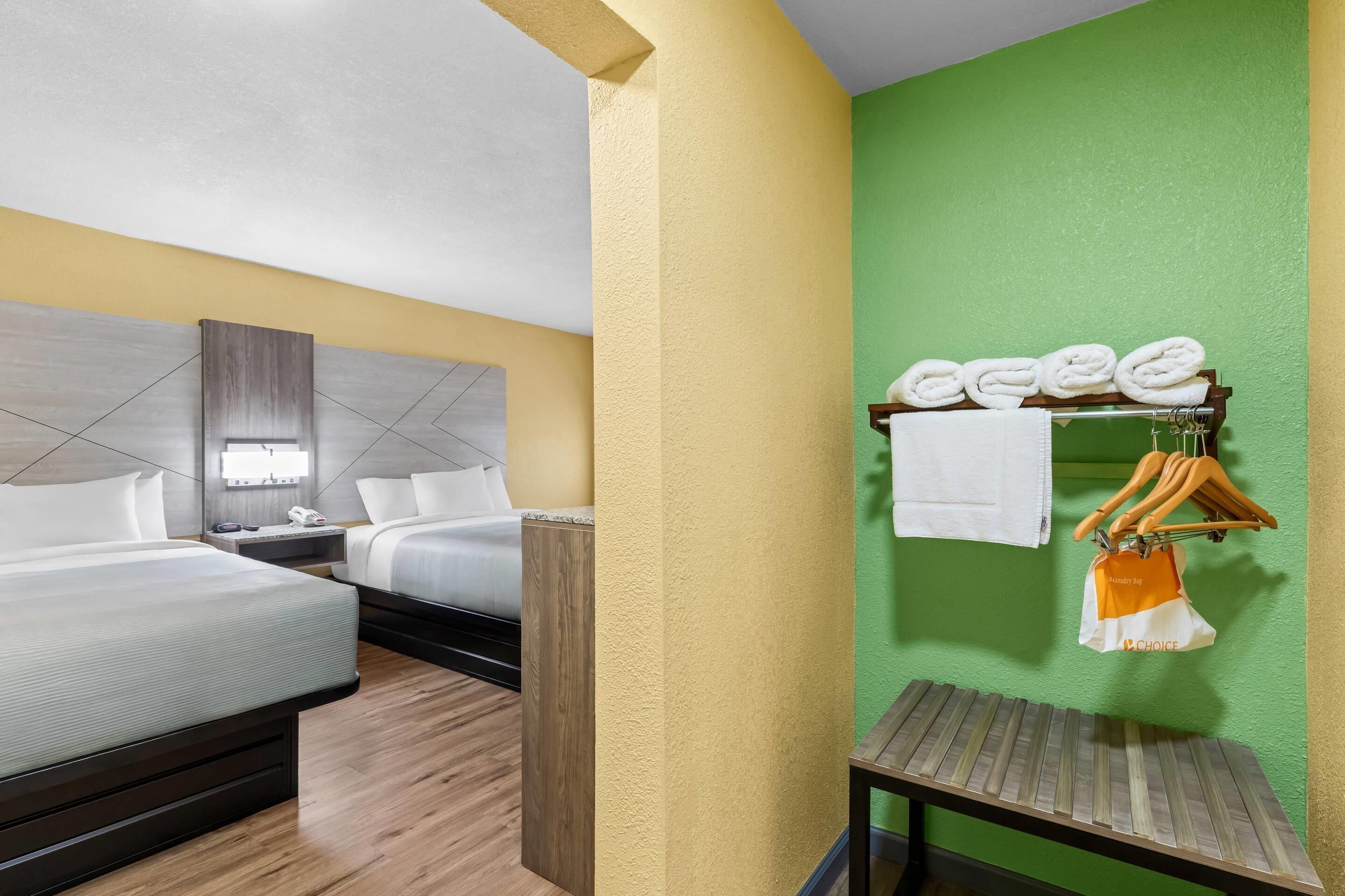 A hotel room with two beds, a wooden headboard, a green accent wall, a small table, and a towel rack with folded towels and a hanging bag.