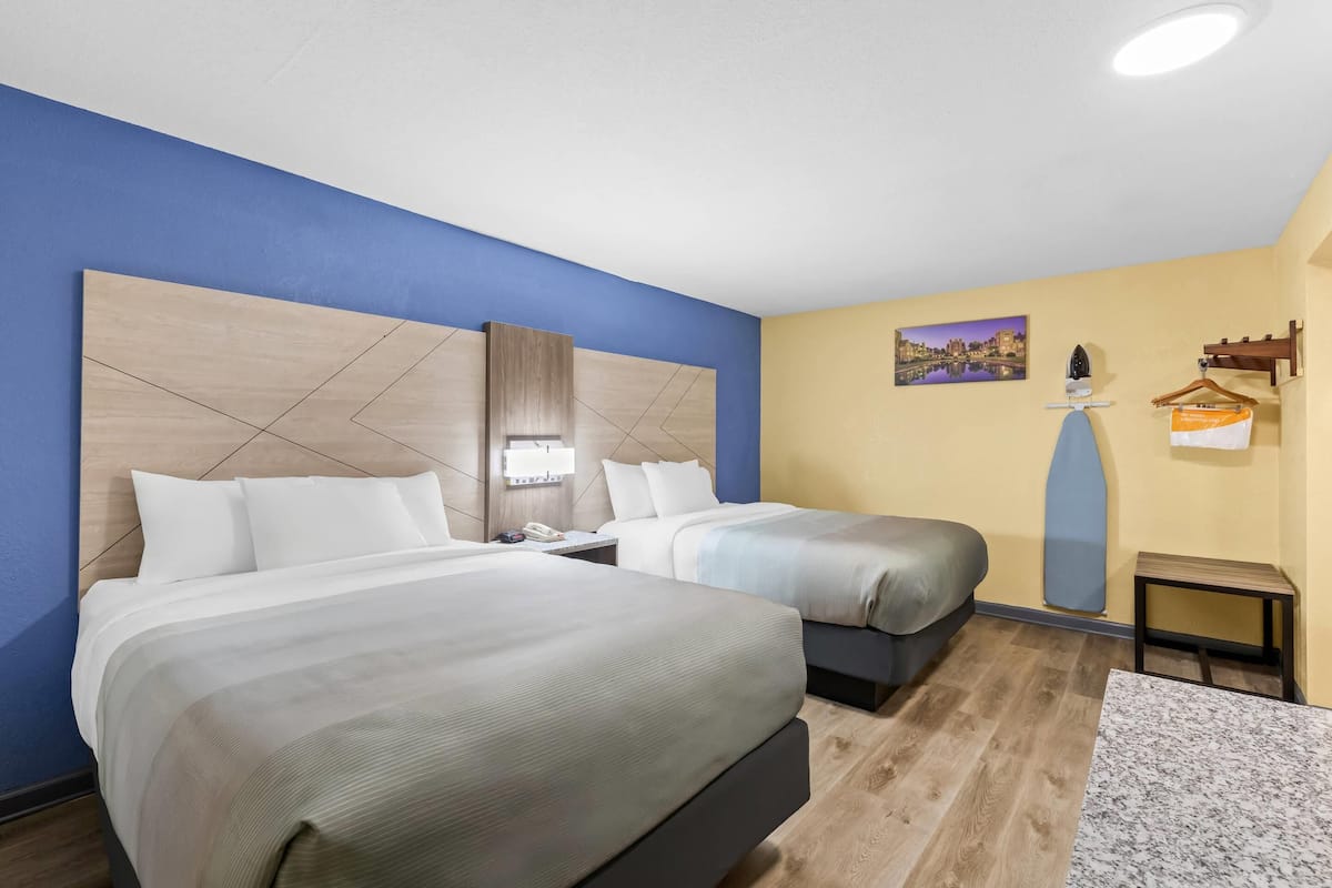 A hotel room with two beds, a nightstand, a lamp, a painting, and a small table.