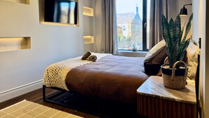 Een moderne hotelkamer met een groot bed, een flatscreen televisie aan de muur, een raam met uitzicht op gebouwen, en een nachtkastje met een plant.