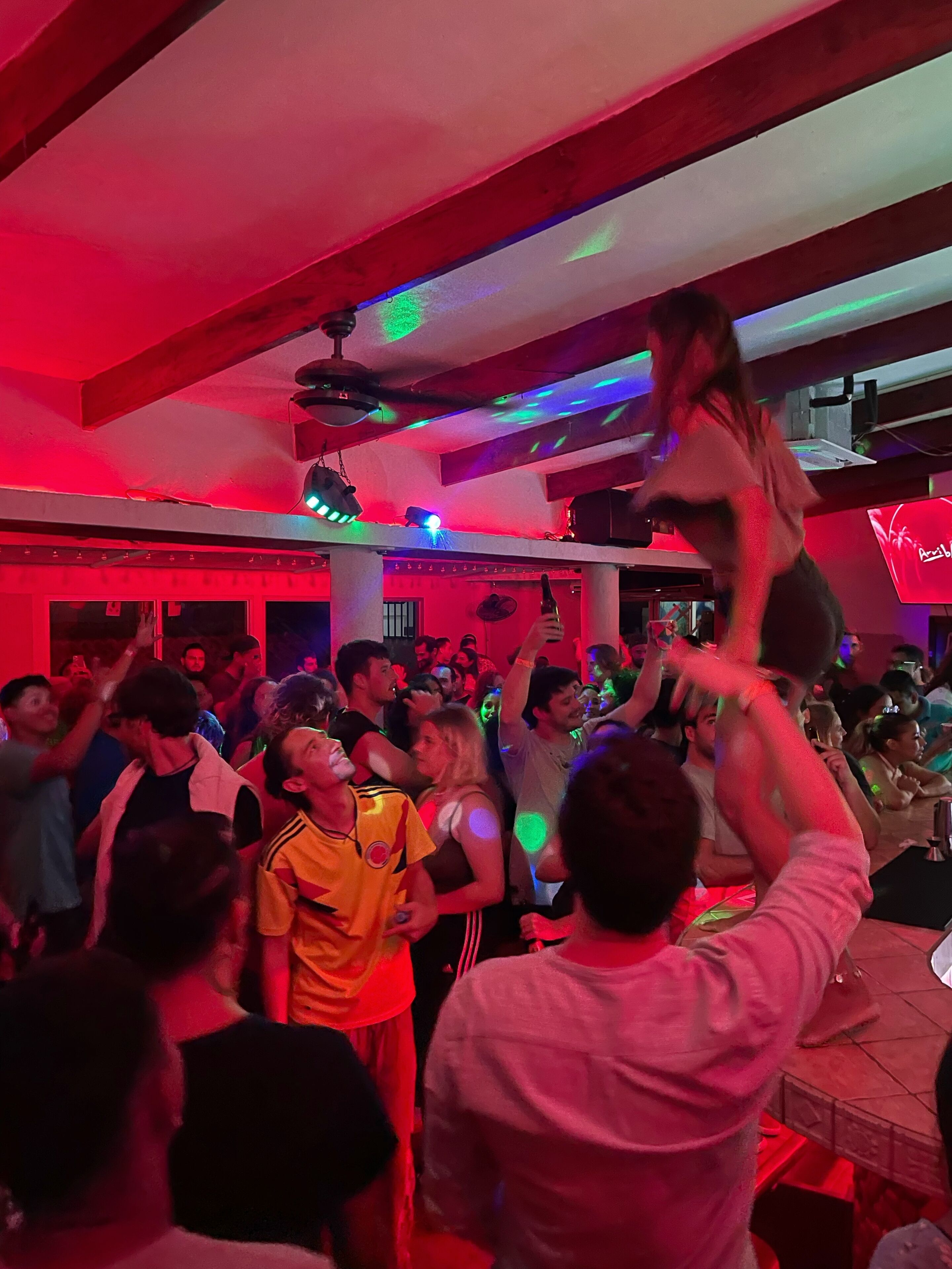 A crowded indoor space with people dancing and a visible ceiling fan.