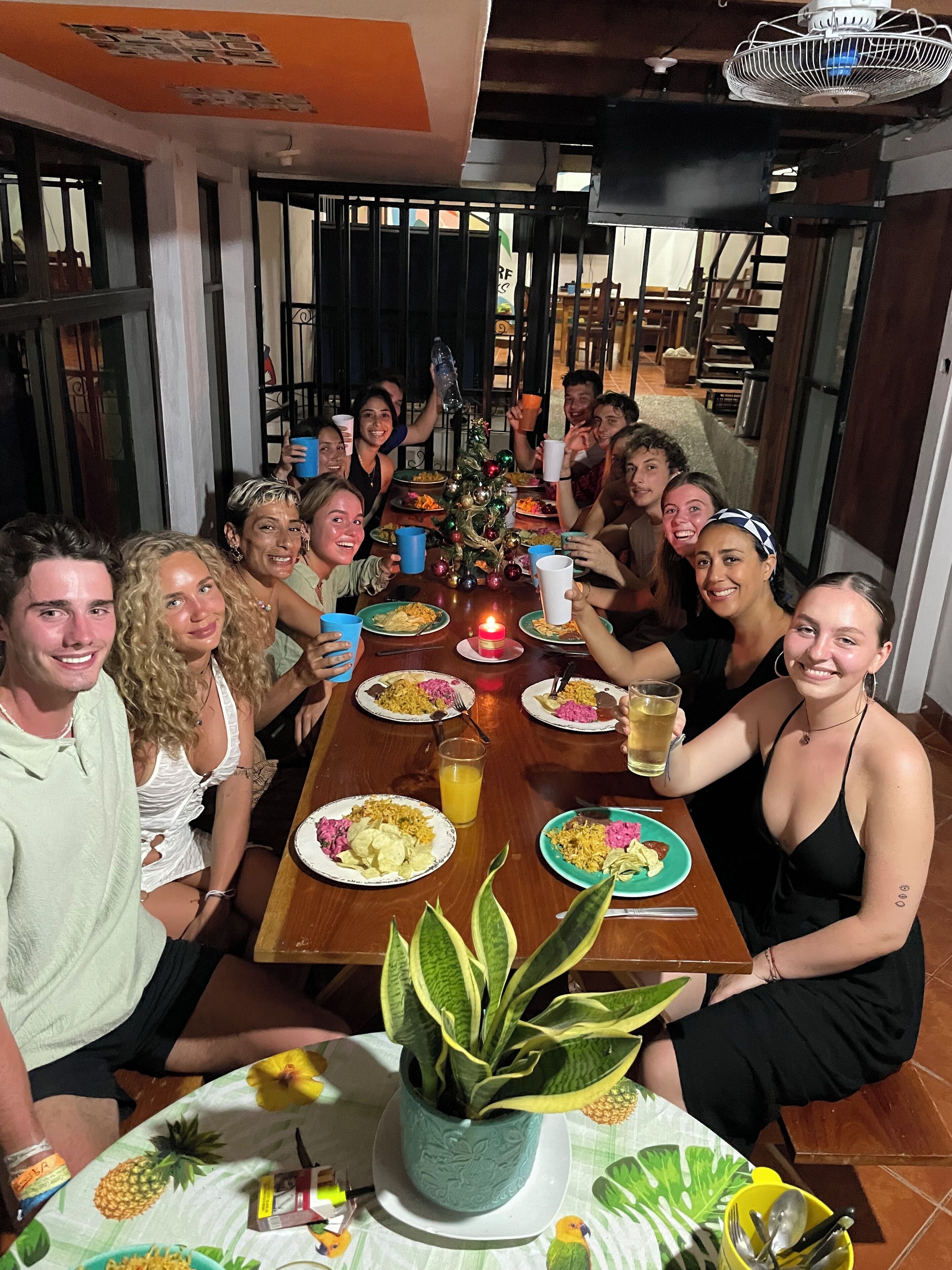 Een groep mensen zit rond een houten eettafel met borden eten, een brandende kaars en een bloempot.