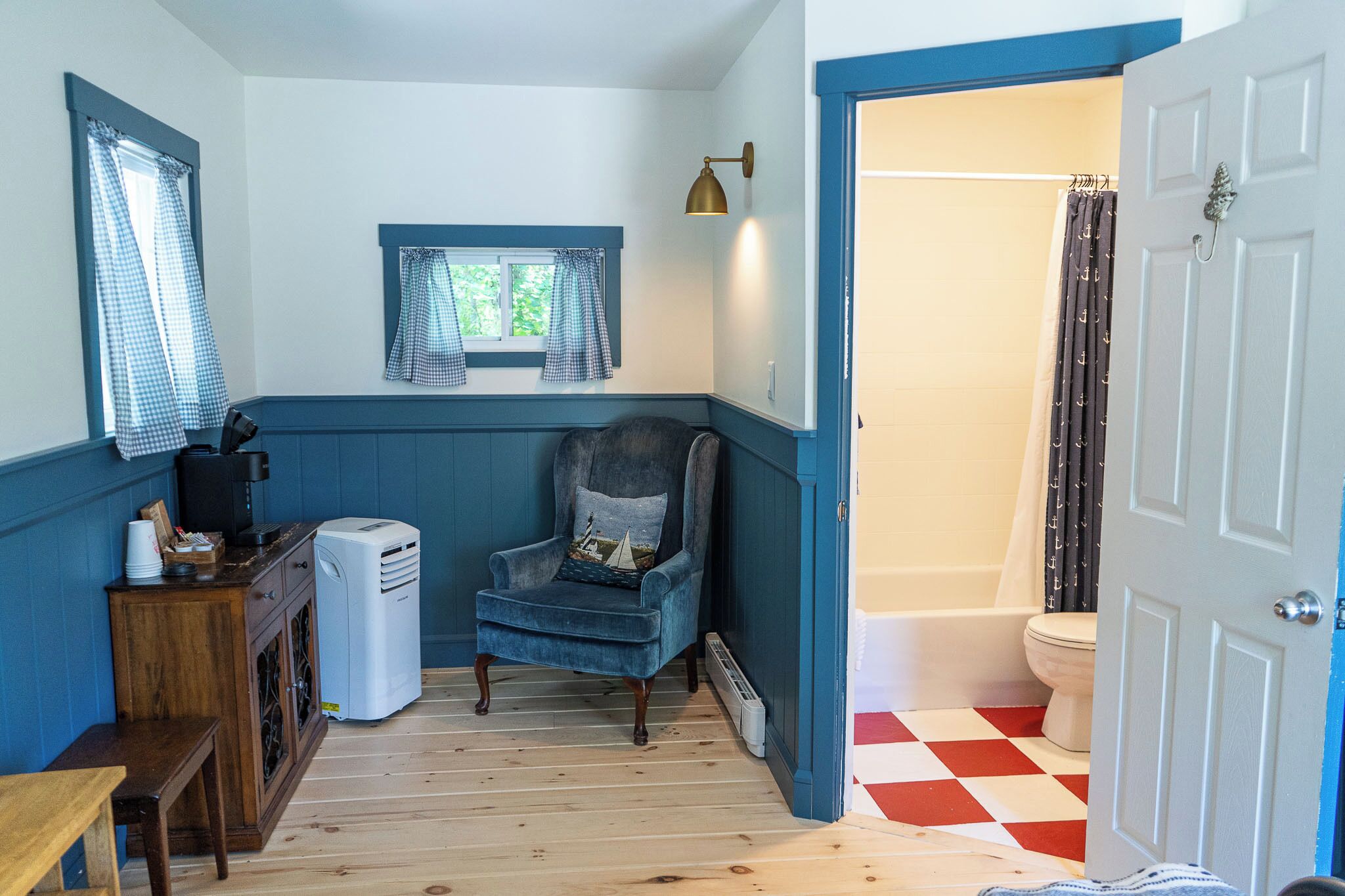 A bathroom with a white toilet, a shower with a black curtain, a blue chair with a pillow, a heater, and a wooden cabinet.