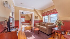 A cozy living room with a wooden floor, a sofa, a coffee table, and a TV. There is a window with curtains and a view of the outside. - Spacious Garden-View King Suite in 150-Year-Old Manor House (Bocabec)