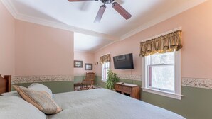 Une chambre à coucher comprenant un lit, une chaise, un bureau, un téléviseur et une fenêtre avec des rideaux.