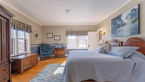 Une chambre avec un grand lit, une commode, une chaise et un bureau. Il y a des fenêtres avec des rideaux, un tableau au mur et une lampe sur le bureau.