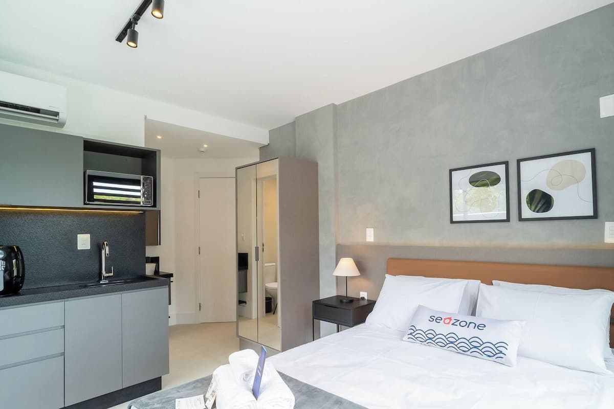 A modern hotel room with a bed, a nightstand, a lamp, a mirror, a wardrobe, and a kitchenette.