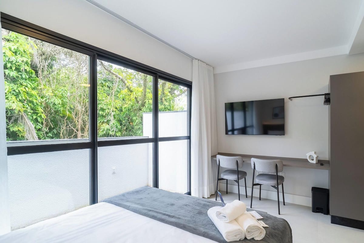 A modern hotel room with a large window, a bed, a TV, and a kitchenette.