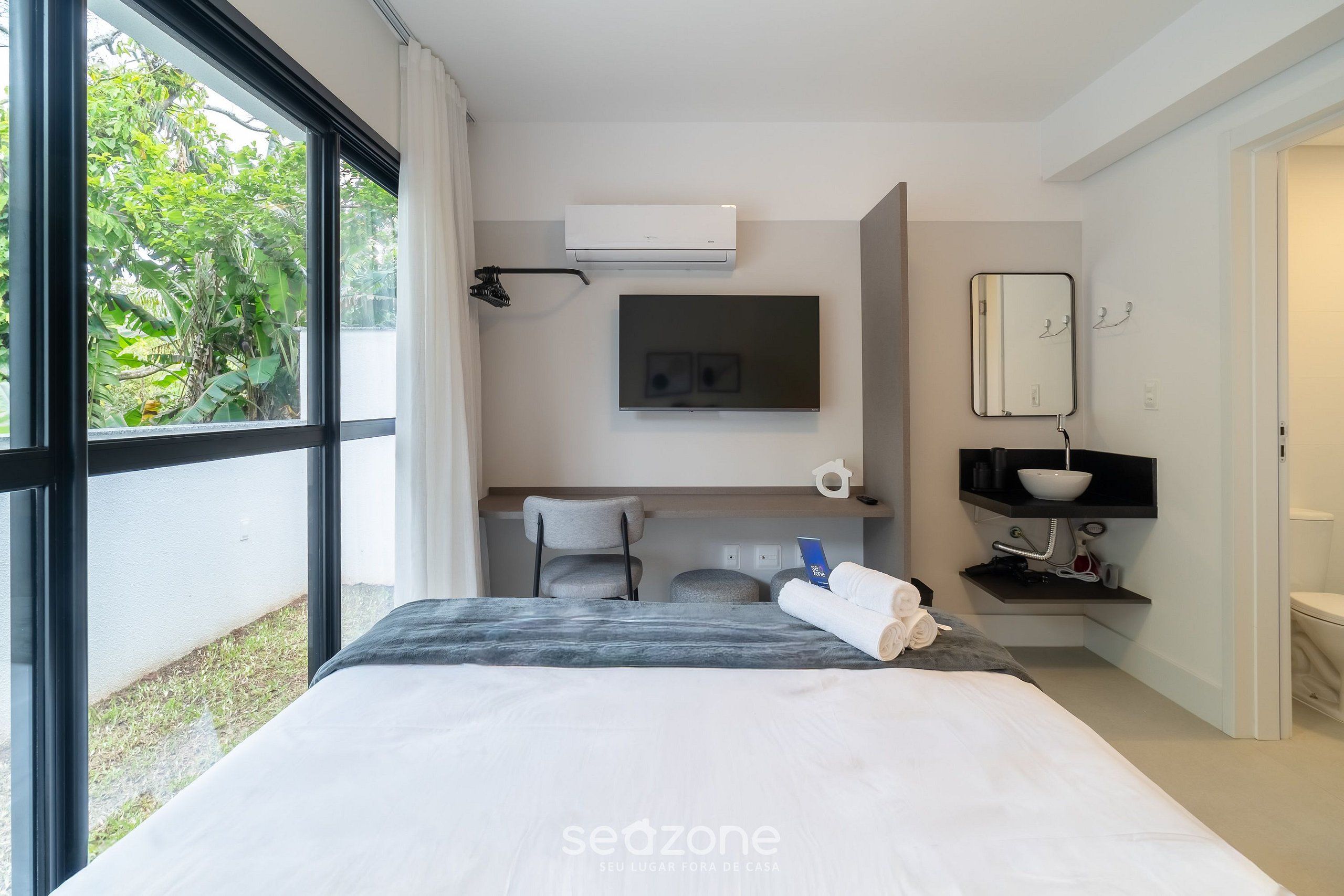A modern hotel room with a bed, a flat-screen TV, a mirror, a sink, and a window with a view of greenery.