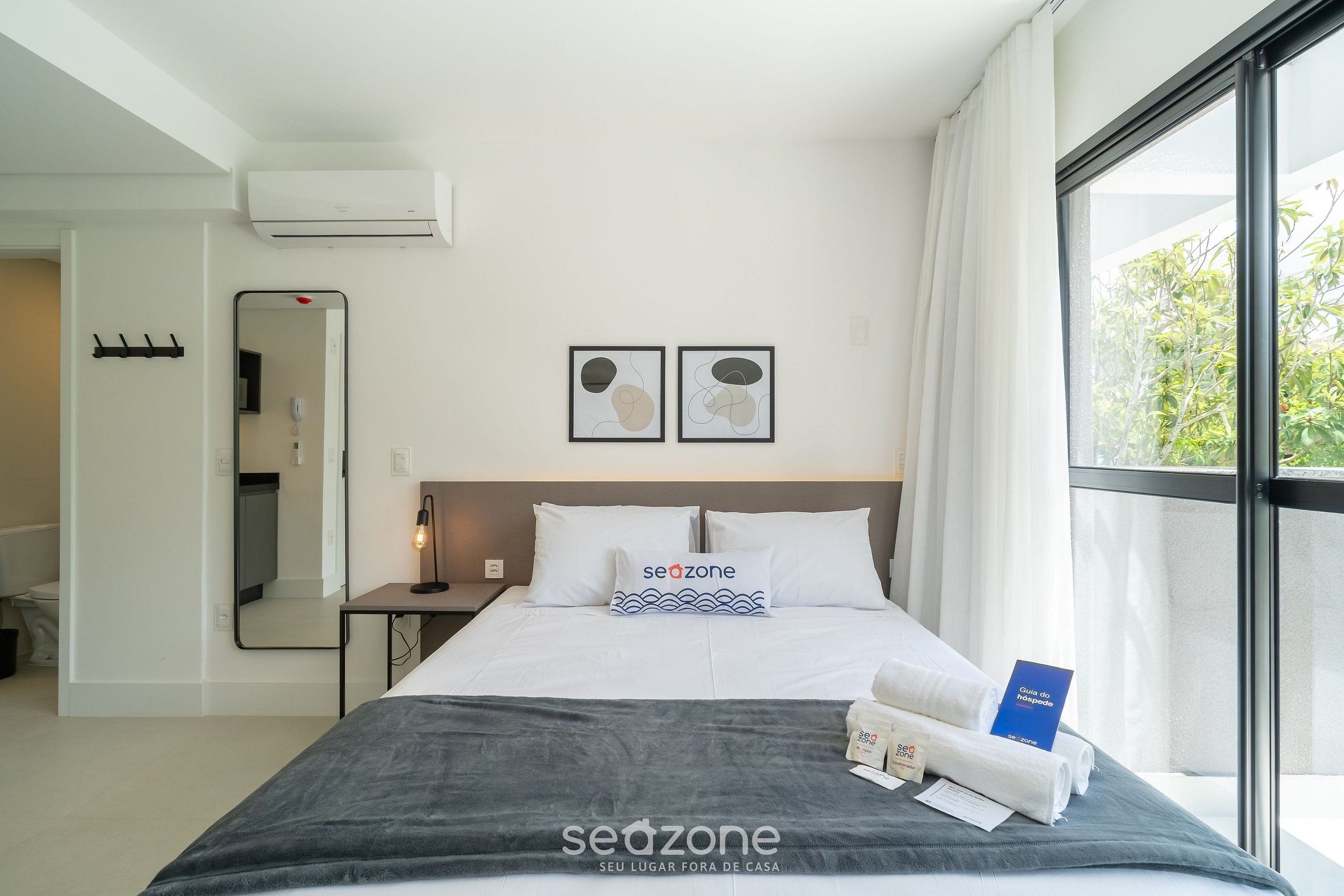A hotel room with a bed, a nightstand, a mirror, two framed artworks, and a window with a view.