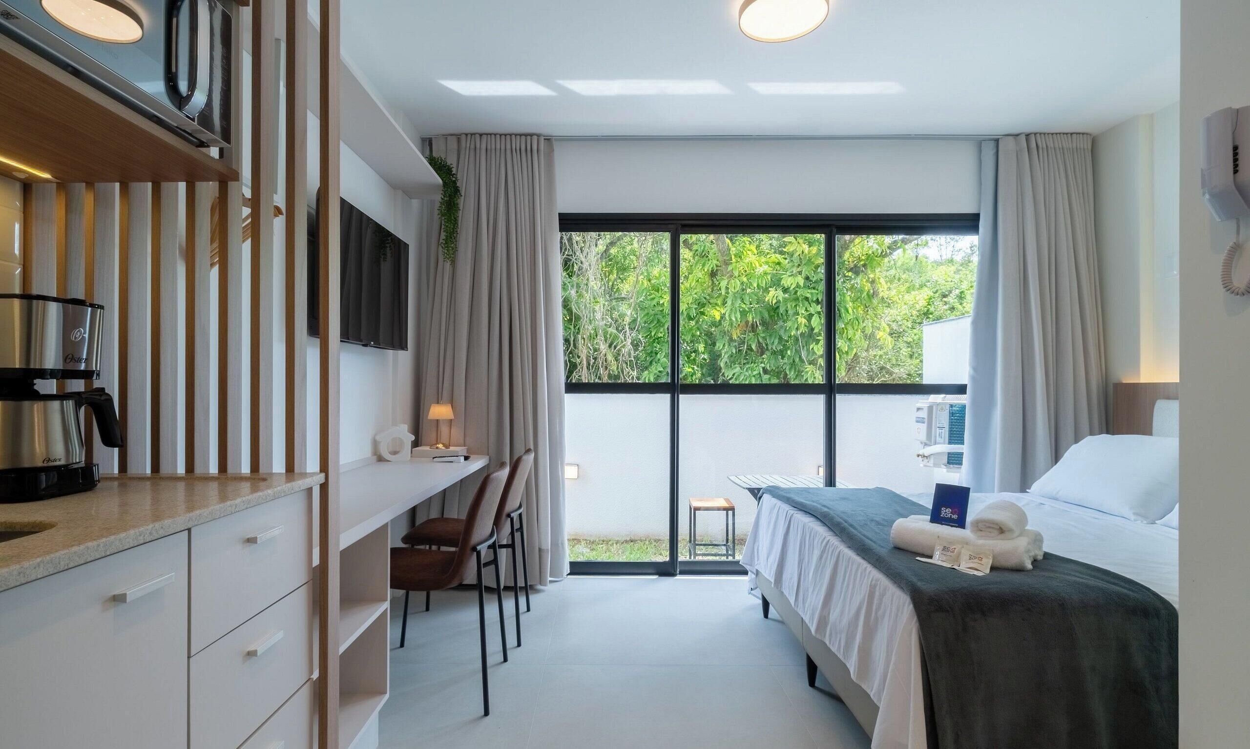 Una habitación de hotel moderna con cama, escritorio, silla, cafetera y un amplio ventanal con vistas al verdor.