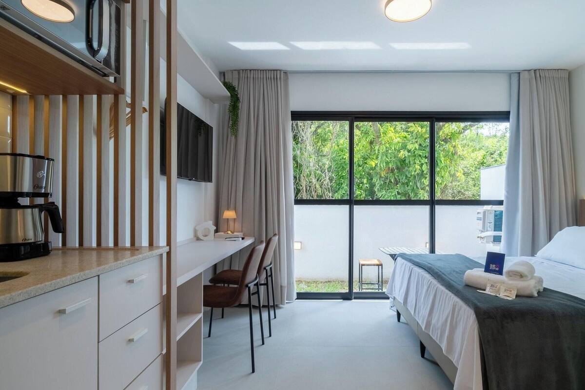 Une chambre d’hôtel moderne équipée d’un lit, d’un bureau, d’une chaise, d’une machine à café et d’une grande fenêtre donnant sur un paysage verdoyant.