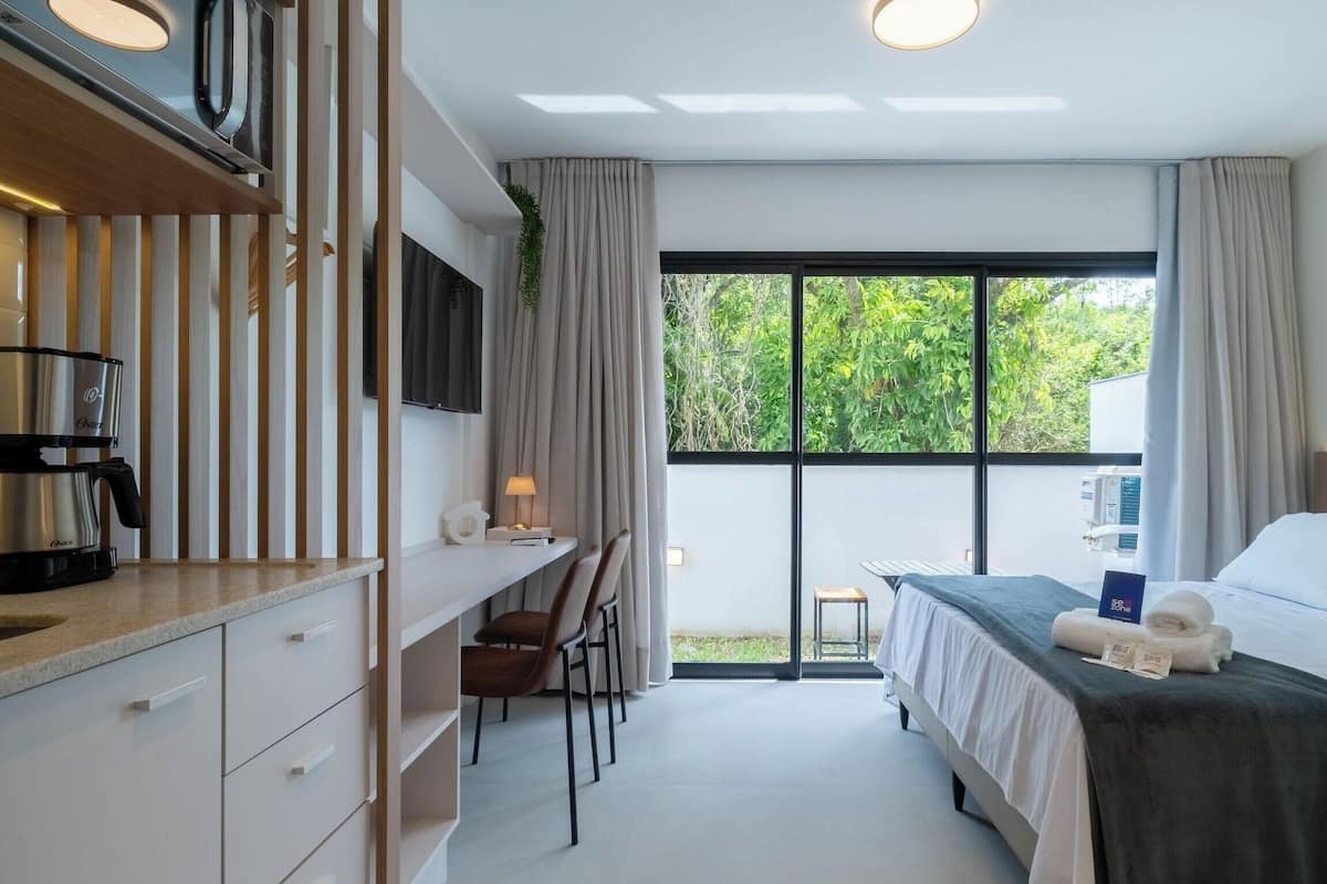 A modern hotel room with a bed, a desk, a chair, a coffee maker, and a large window with a view of greenery.