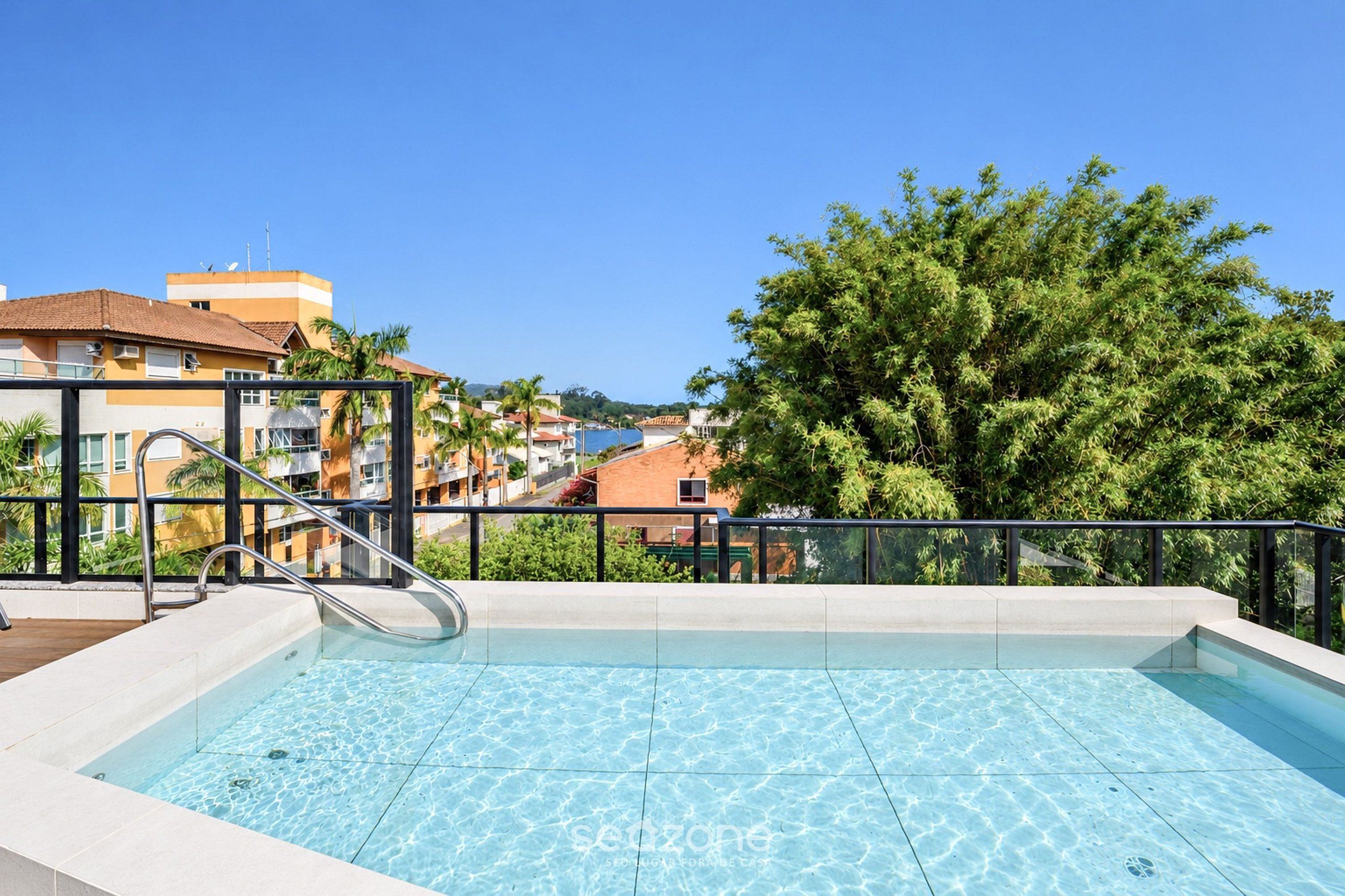 Una piscina amb aigua cristal·lina, envoltada d'una barana metàl·lica, amb vista a una zona residencial amb edificis i arbres.