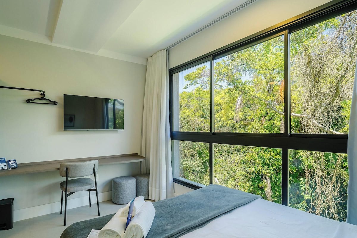 A modern hotel room with a large window, a flat-screen TV, a desk, a chair, and a bed.