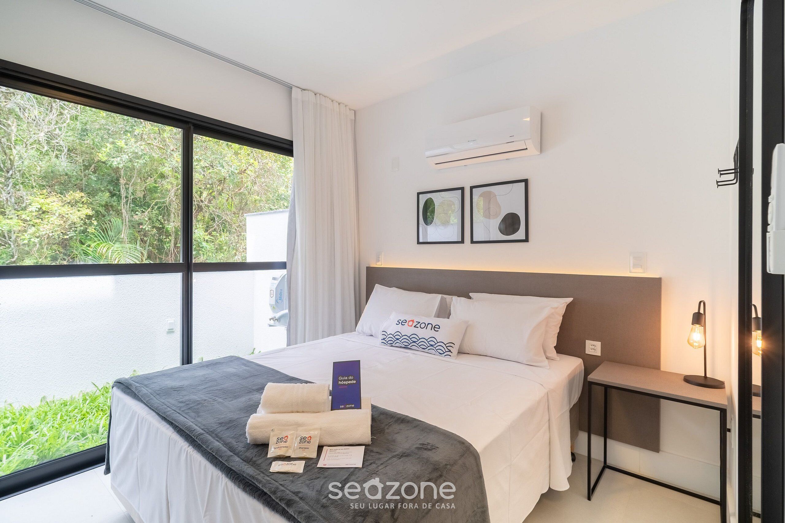 Habitación de hotel moderna con un ventanal grande, una cama con edredón y almohadas blancas, una mesita de noche con lámpara y vistas a zonas verdes.