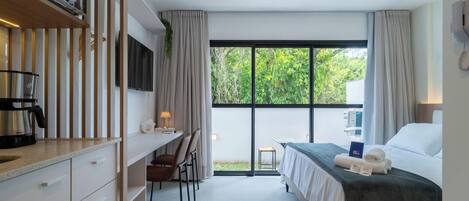 Une chambre d’hôtel moderne avec un lit, un bureau et une chaise. On y trouve une fenêtre avec des rideaux, un téléviseur et une machine à café posée sur le comptoir.