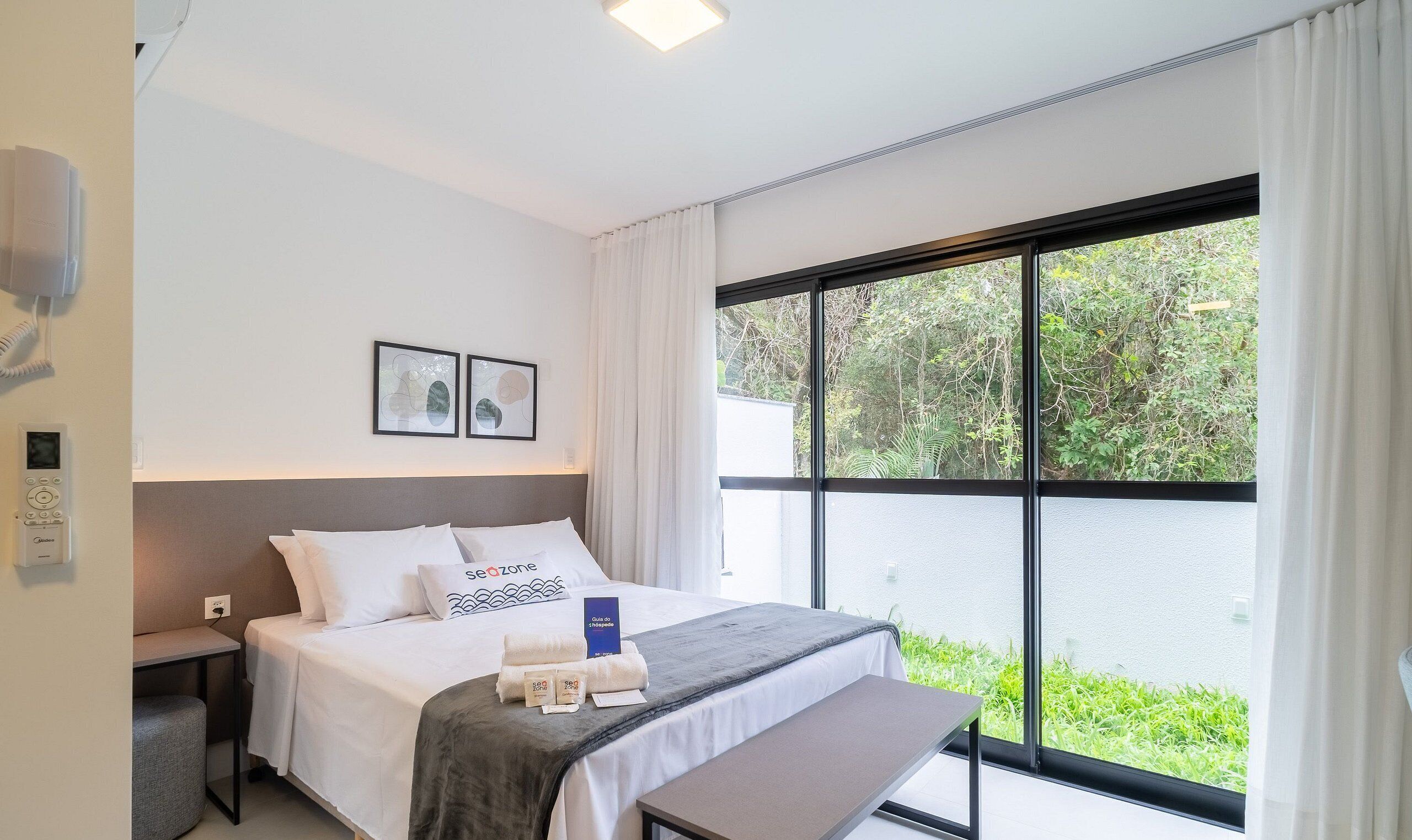 Una habitación de hotel moderna con un ventanal grande, una cama con ropa blanca, una mesita de noche y un panel de control en la pared.