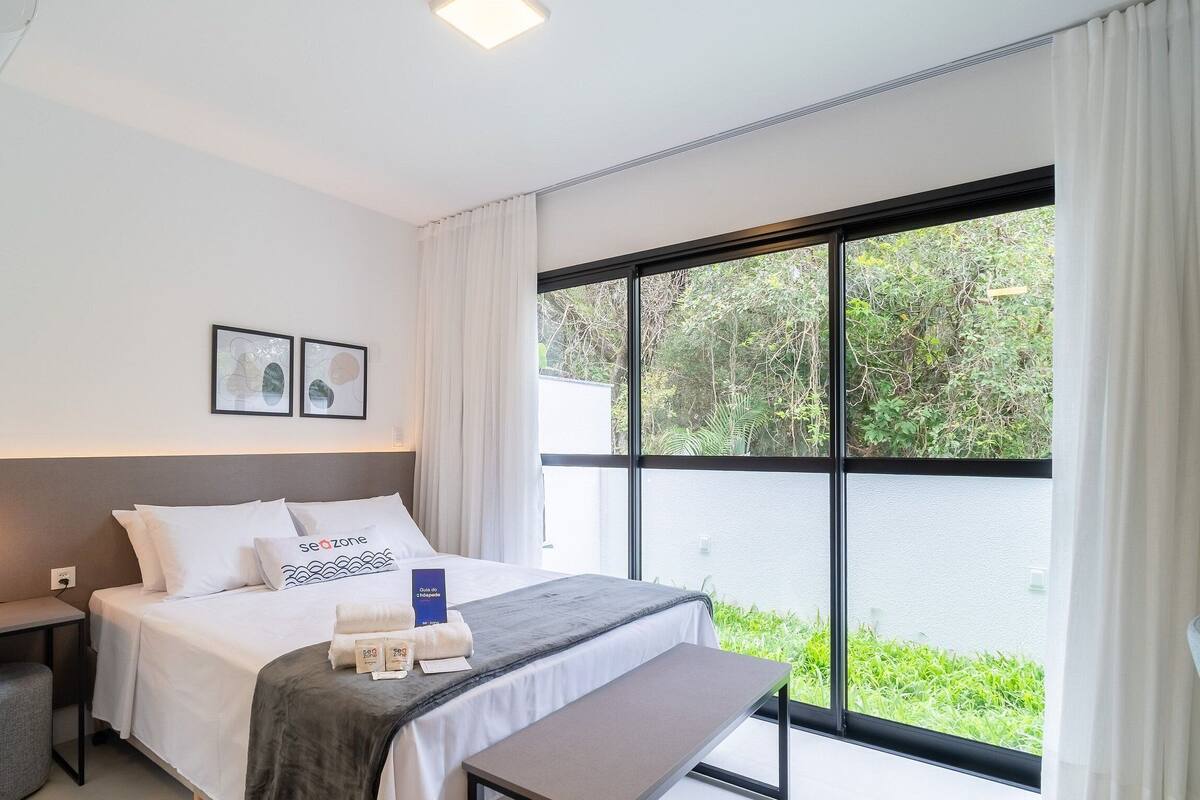 Une chambre d’hôtel moderne dotée d’une grande fenêtre, d’un lit avec une literie blanche, d’une table de chevet et d’un panneau de commande fixé au mur.