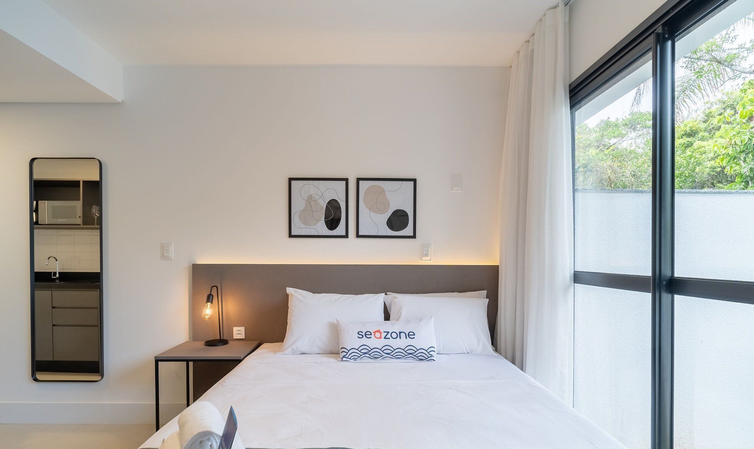Habitación de hotel con cama, mesita de noche, espejo en la pared, ventana con cortinas y dos obras de arte abstractas en la pared.