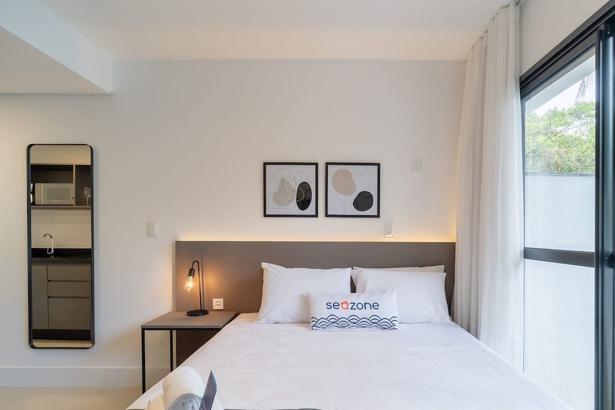 A hotel room with a bed, a nightstand, a wall-mounted mirror, a window with curtains, and two abstract wall art pieces.