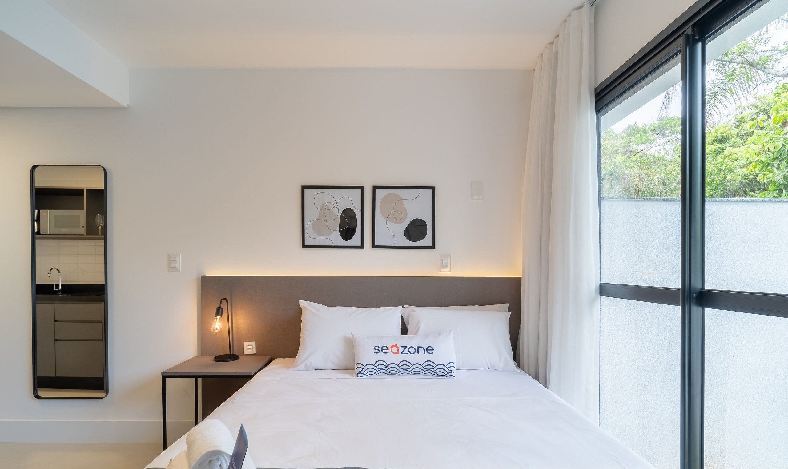 Hotellihuone, jossa on sänky, yöpöytä, seinälle kiinnitetty peili, ikkuna verhoilla sekä kaksi abstraktia taideteosta seinällä.