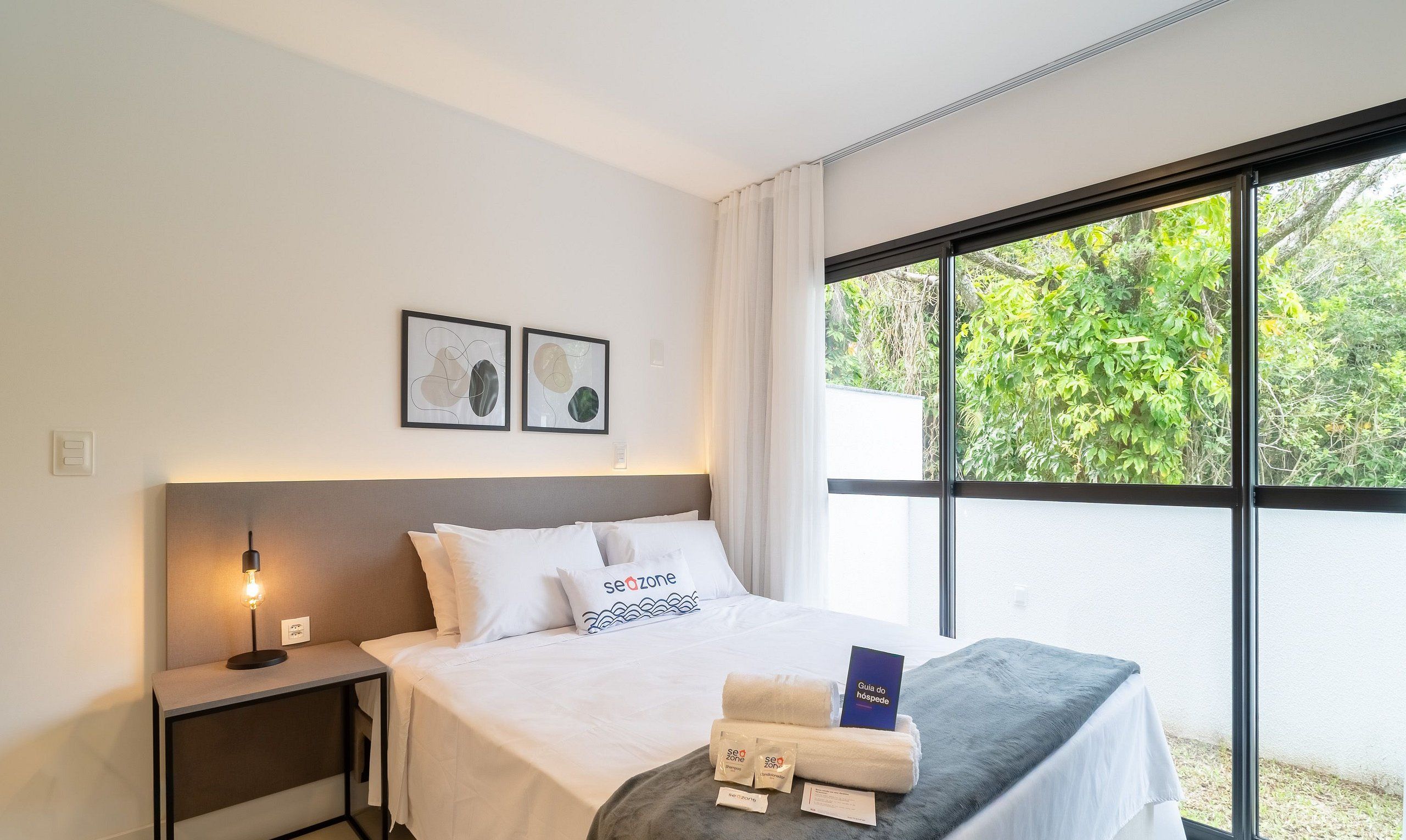 Une chambre d’hôtel avec un lit, une table de chevet avec une lampe, une fenêtre donnant sur un paysage verdoyant et deux tableaux encadrés au mur.
