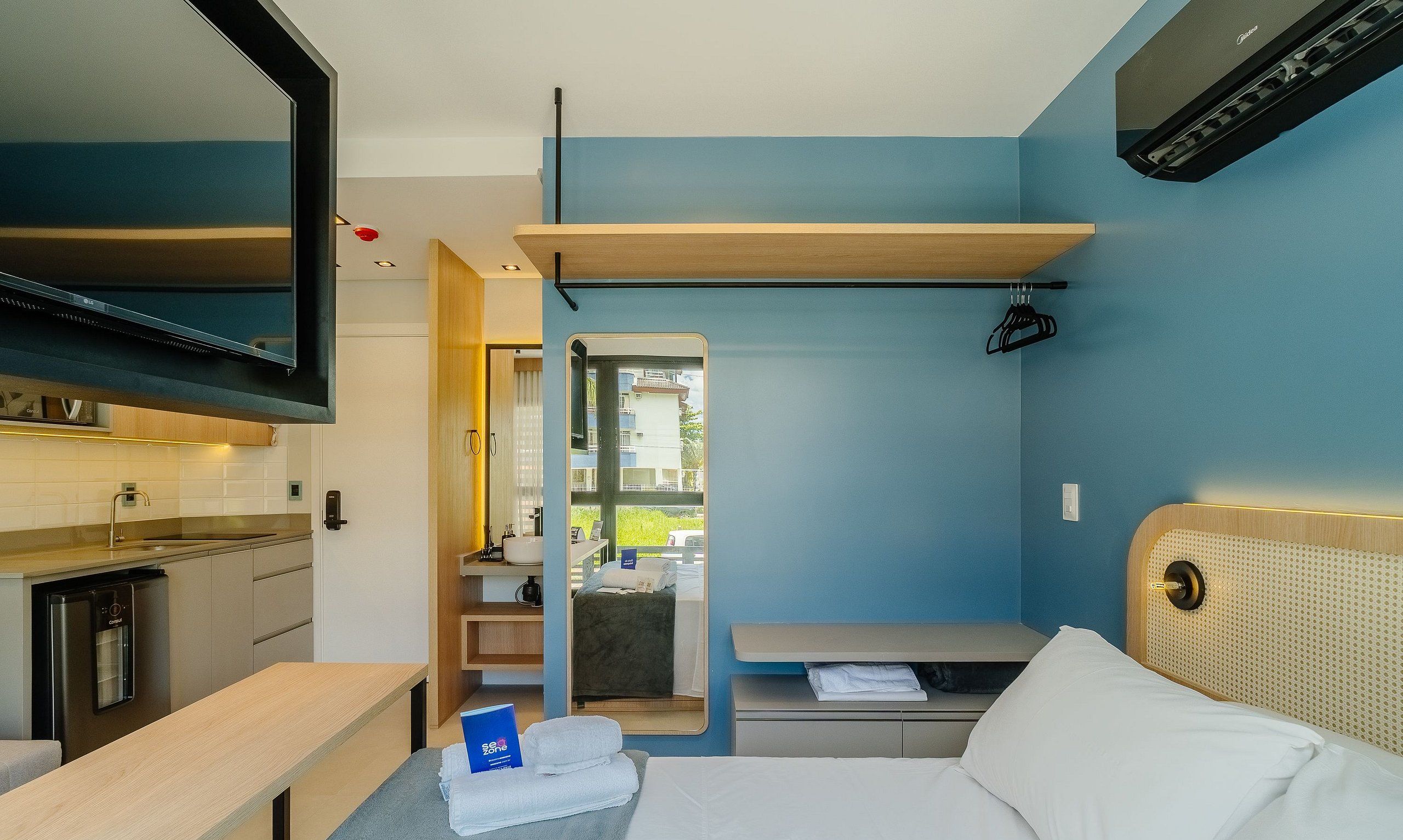 A compact hotel room with a kitchenette, a bed, a bedside table, a mirror, and a window with a view of the outside.