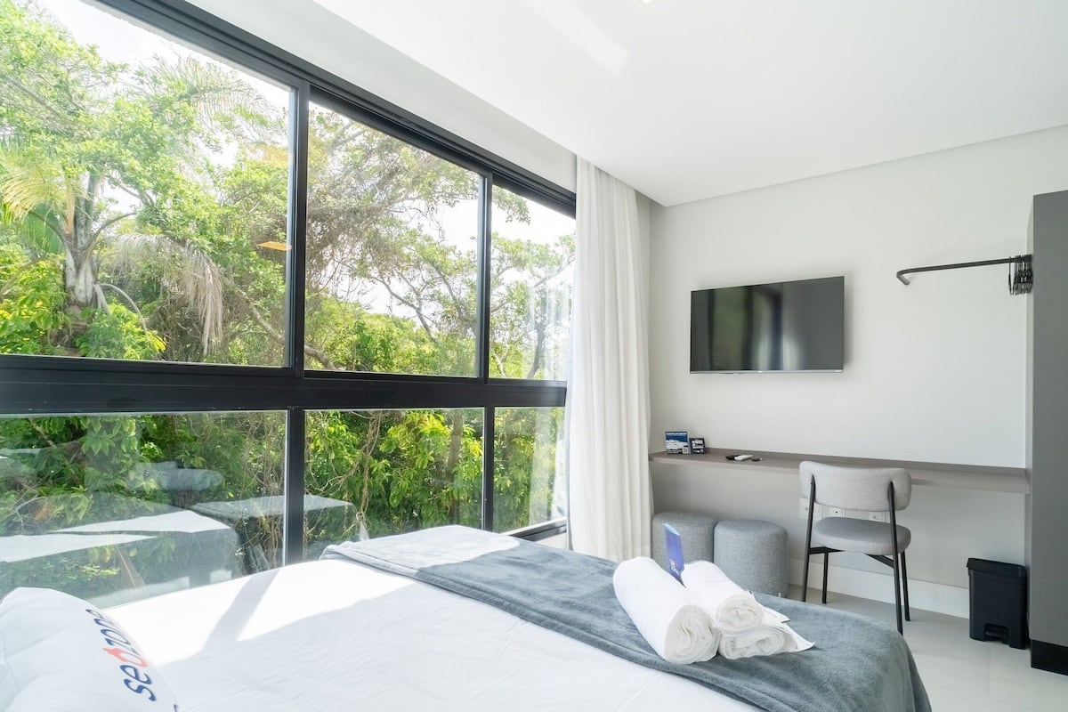 A hotel room with a bed, a desk, a chair, a TV, and a view of greenery outside.