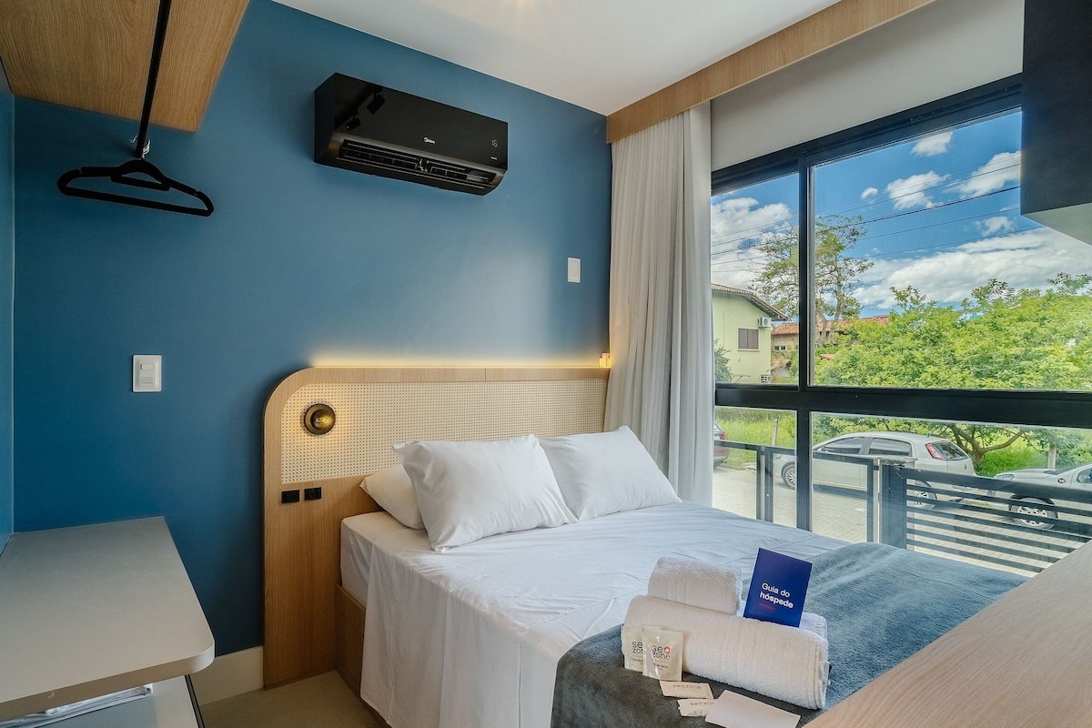 A hotel room with a bed, a desk, a hanging light, a window with a view, and an air conditioning unit.
