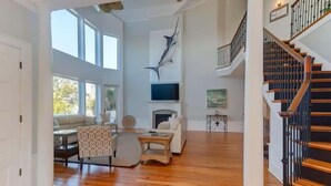 Uma sala de estar ampla com piso de madeira, lareira, TV fixada na parede e escada com corrimão de madeira.