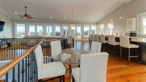 Uma ampla área de refeições com uma mesa de vidro cercada por cadeiras brancas, uma escada de madeira e um espaço de cozinha com banquetas.