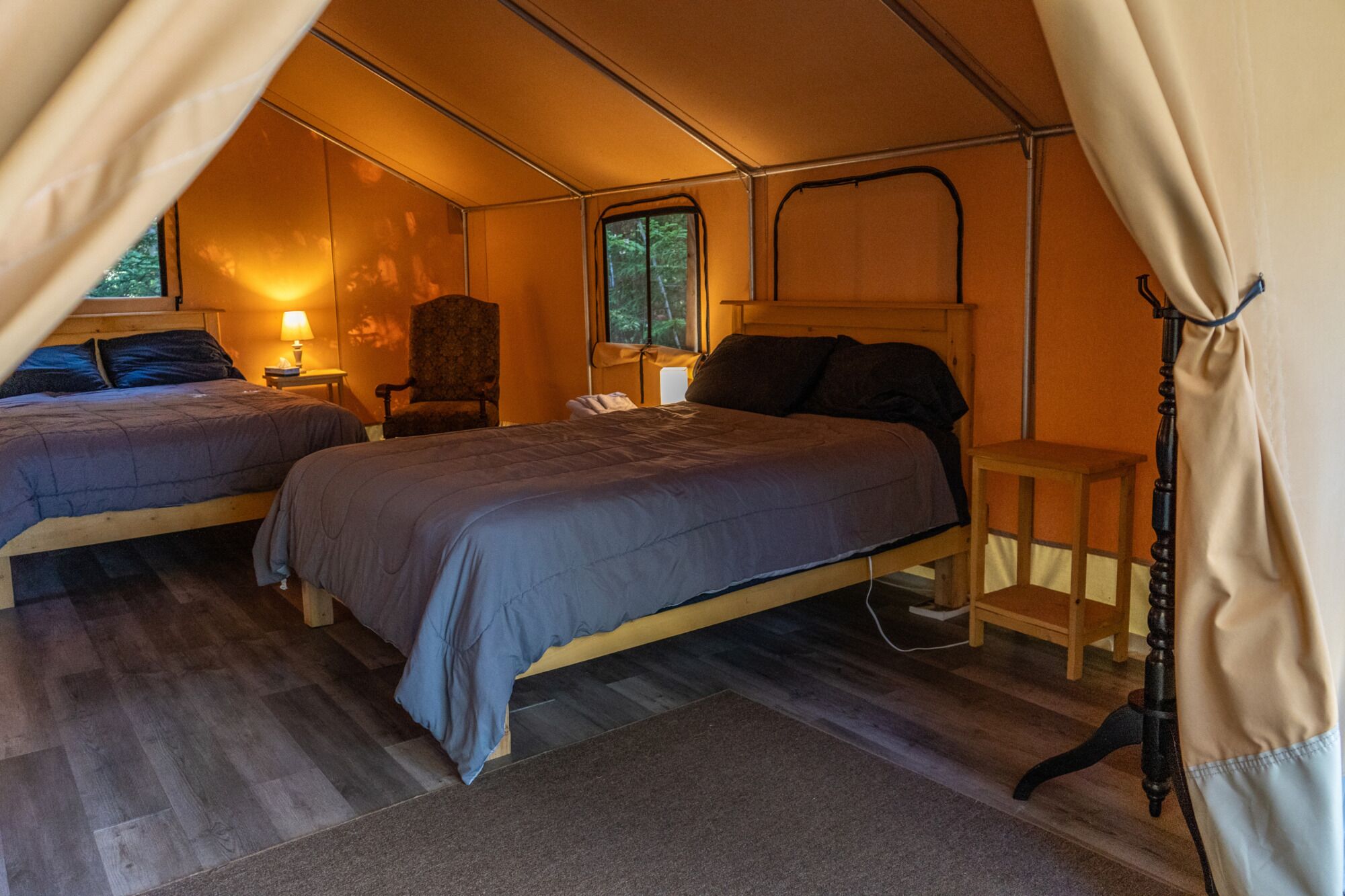 A tented accommodation with two beds, a chair, and a bedside table.