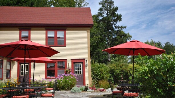 Un bâtiment doté d’un toit rouge et d’un revêtement beige, avec un espace extérieur aménagé avec des parasols rouges et un sol en pierre.
