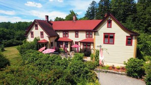 Une grande maison à deux étages, avec un toit rouge et de nombreuses fenêtres à cadre rouge. On y trouve des parasols d’extérieur et un chemin de pierre menant à l’entrée.