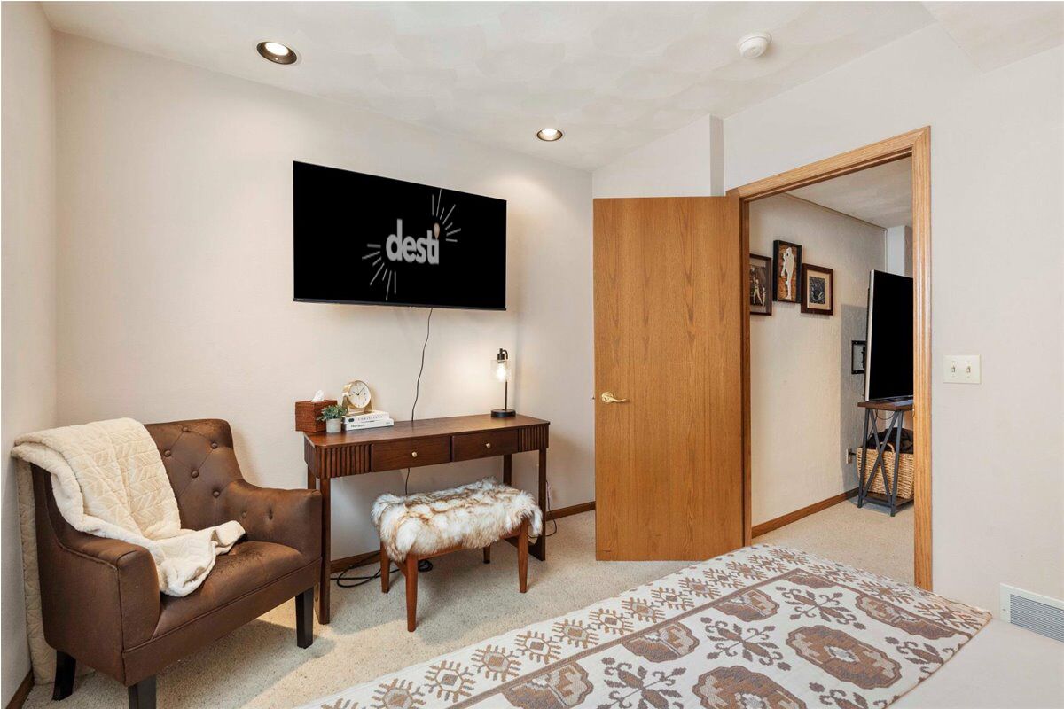 Una sala de estar con un sofá de cuero marrón, un escritorio de madera, un tapete estampado y un televisor montado en la pared.