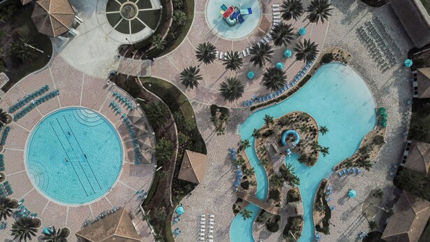 Vista aerea di un resort con una grande piscina centrale, una piscina circolare più piccola e uno scivolo d'acqua.