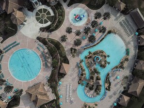 Vista aérea de un complejo turístico con una amplia piscina central, una piscina circular más pequeña y un tobogán de agua.