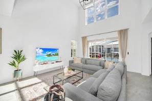 A spacious living room with a large gray sofa, a coffee table, and a TV displaying a tropical beach scene. - Champions Luxe - Resort & Arcade - Near Clubhouse! (Davenport)
