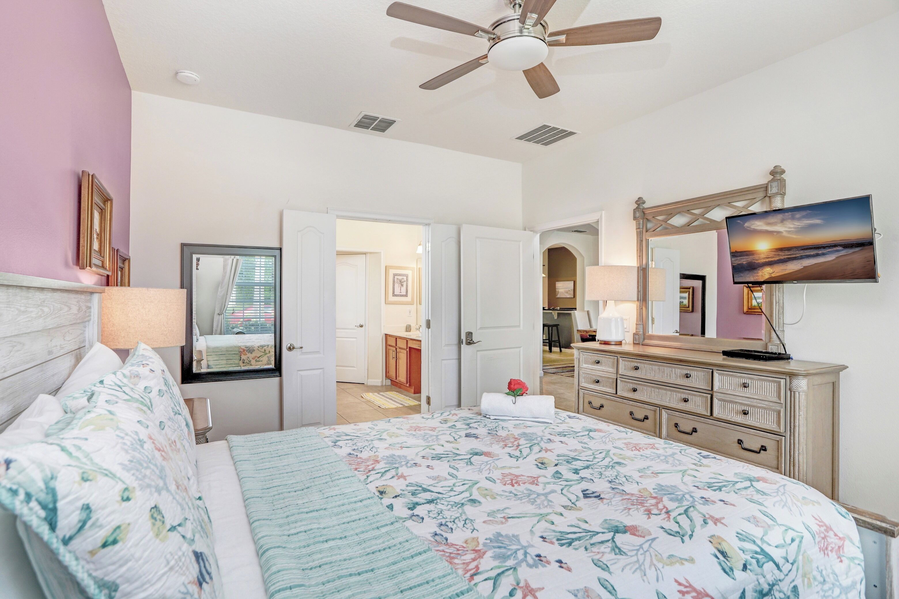 A bedroom with a bed, a ceiling fan, a dresser, a TV, and a bathroom visible through an open door.