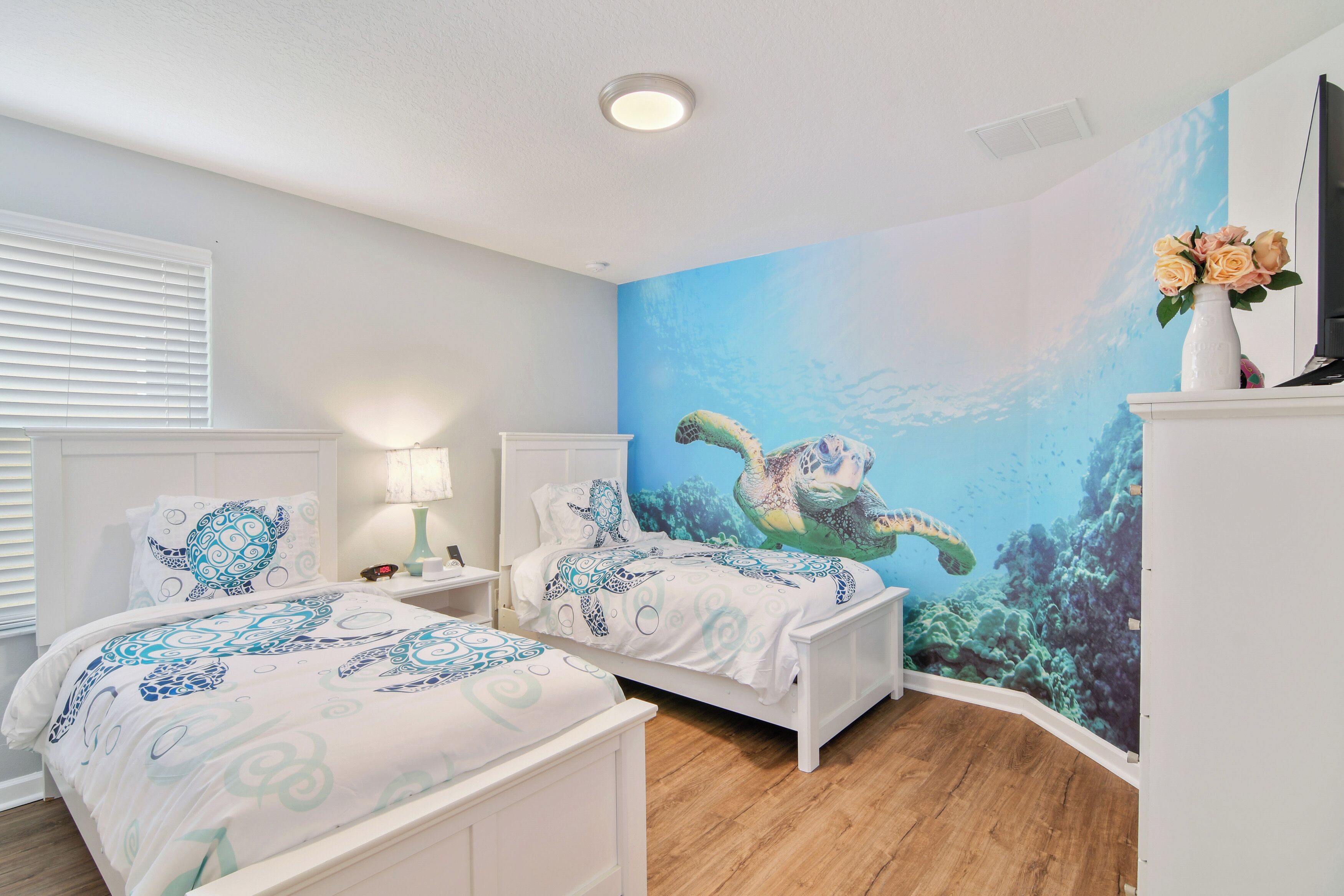 A bedroom with two beds, a mural of a sea turtle, and a white dresser with a vase of flowers.