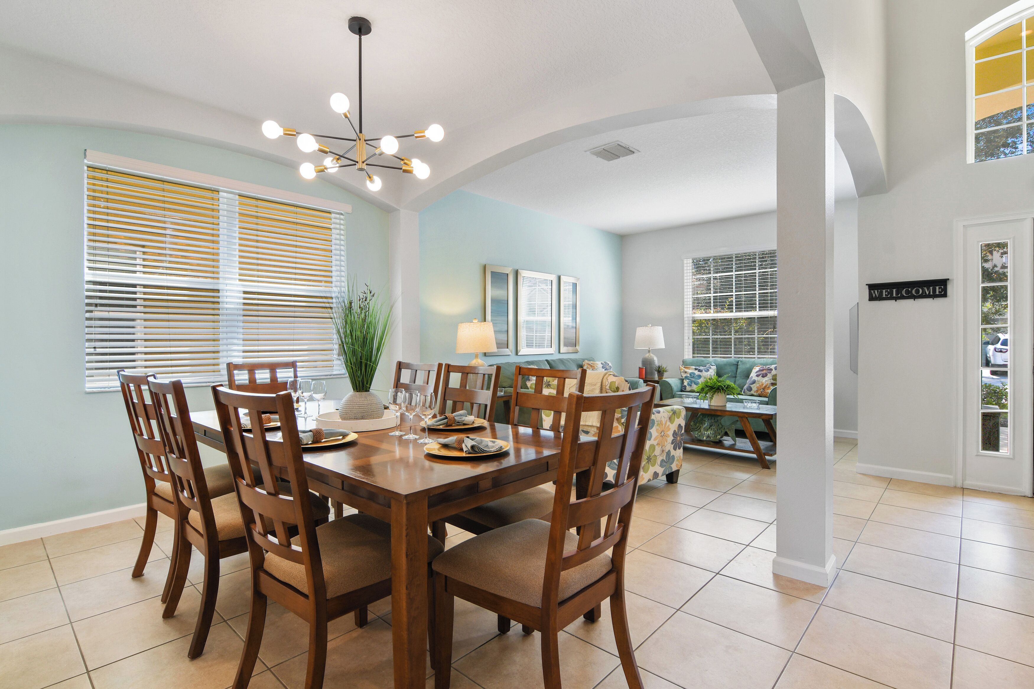 A dining area with a wooden table and chairs, a chandelier, and a view into a living room with a sofa and a 'WELCOME' sign.