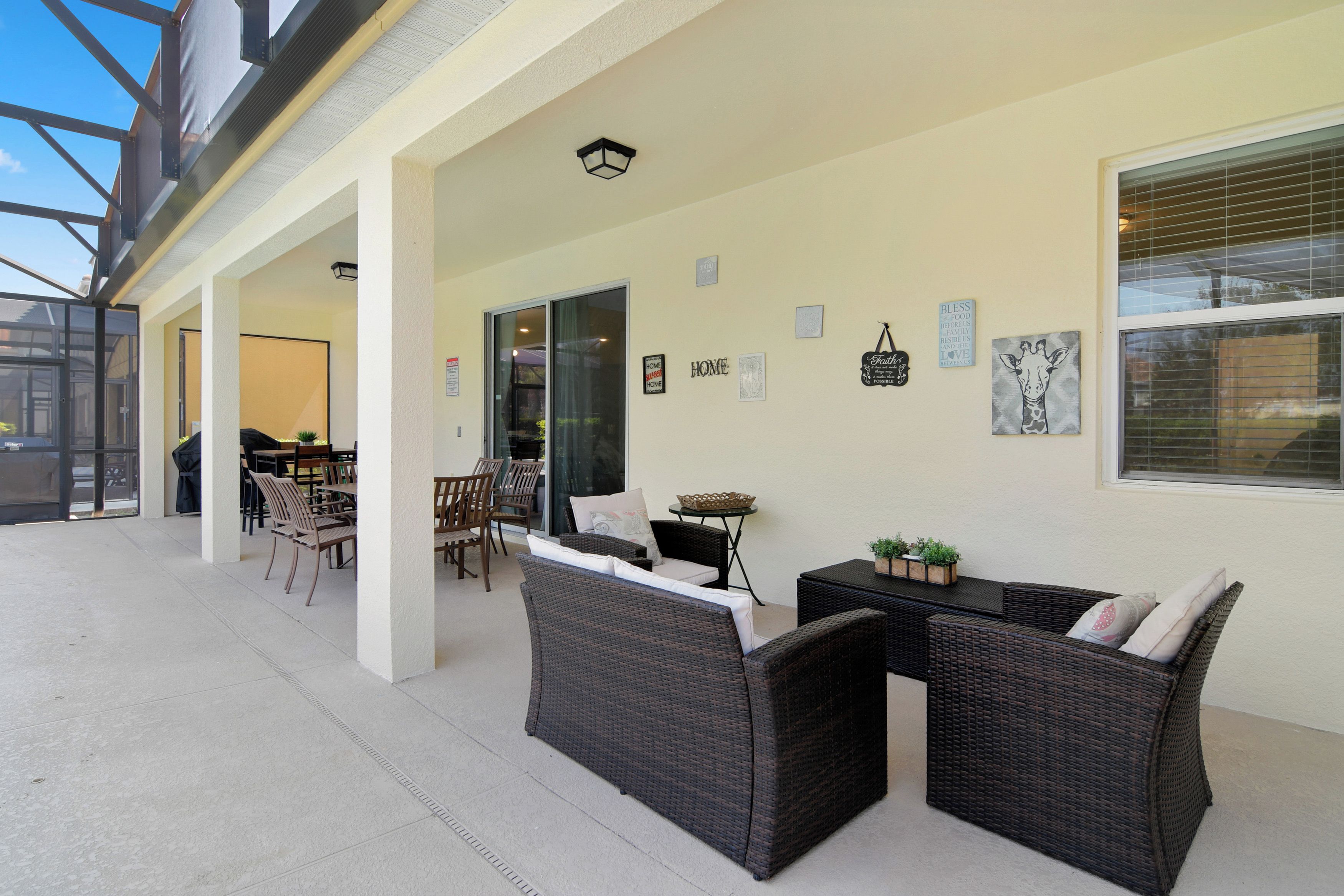 An outdoor patio area with wicker furniture, a dining table, and chairs. There are wall decorations and a window with blinds.