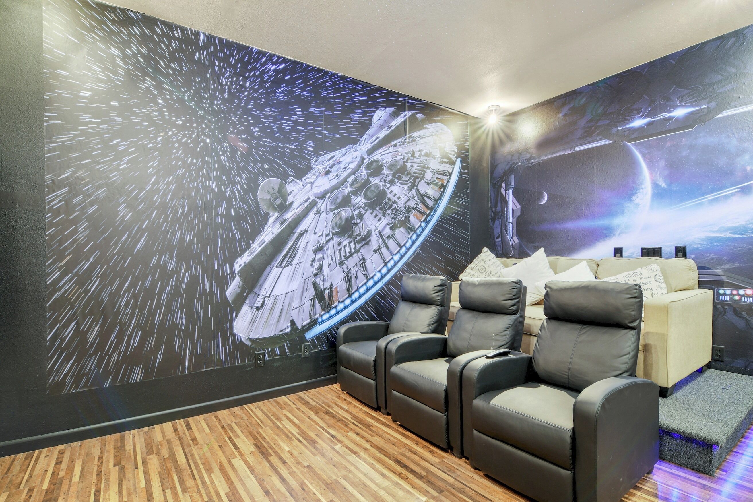 A home theater room with a large screen displaying a space-themed movie, three reclining chairs, and a wooden floor.