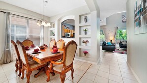 Uma área de refeições com uma mesa e cadeiras de madeira, uma cozinha ao fundo e uma sala de estar com um sofá e abajur.
