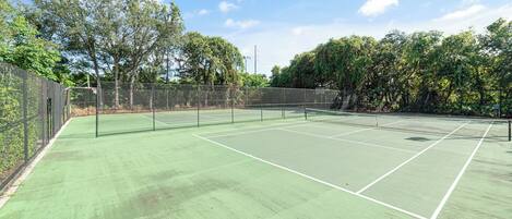 Una cancha de tenis con superficie verde, rodeada por una valla y árboles.