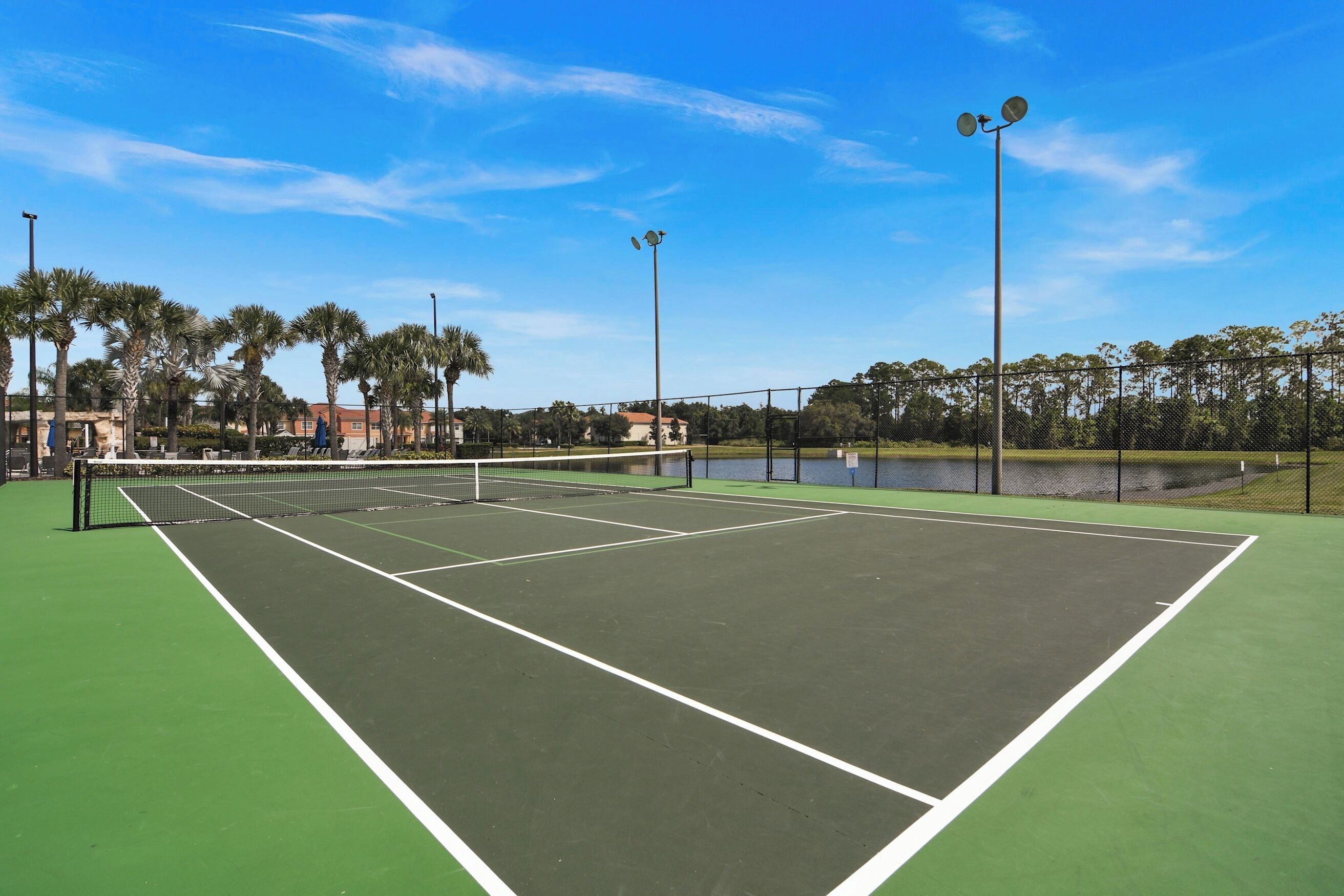 Un court de tennis avec un filet, entouré d’une clôture, de palmiers et d’une étendue d’eau.