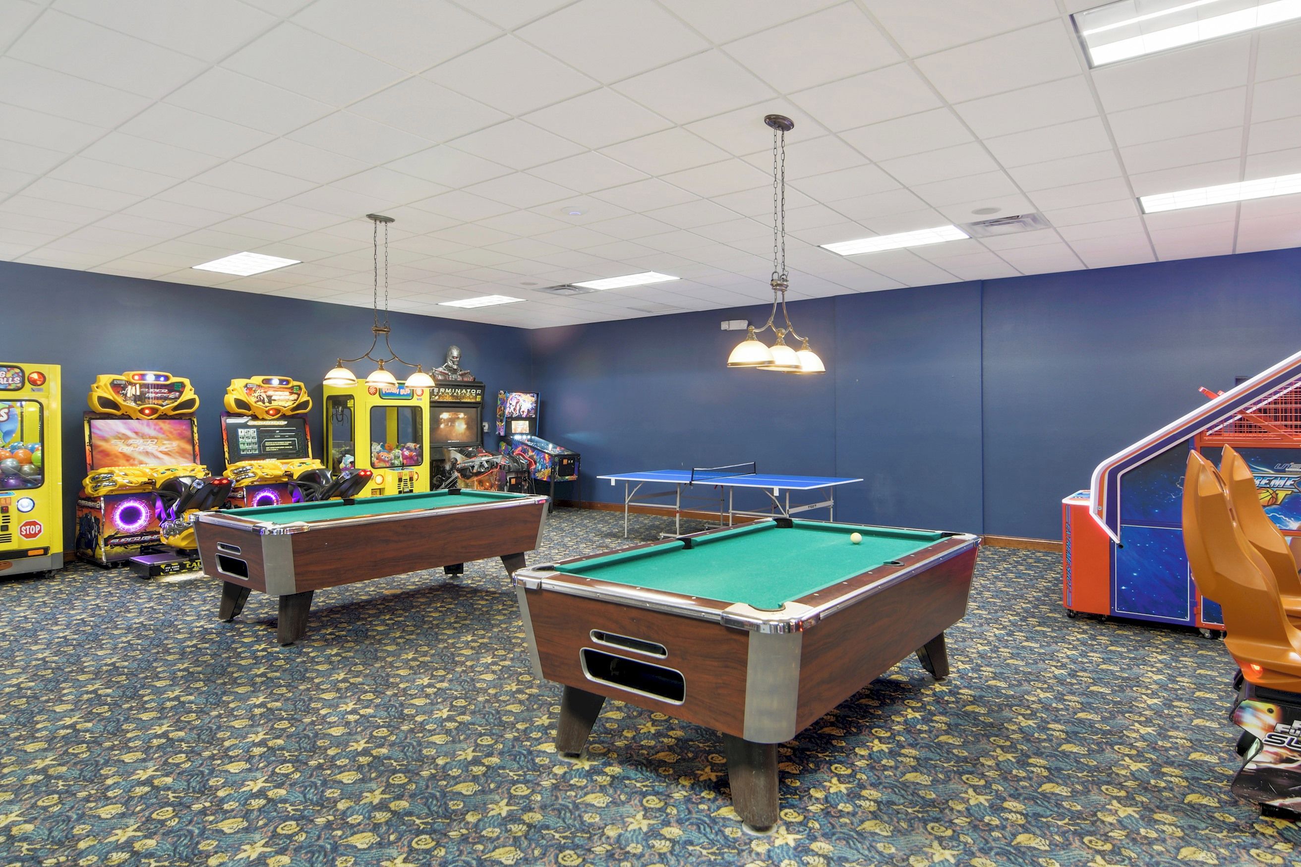 Une table de billard, deux bornes d’arcade et une table de ping-pong se trouvent dans une pièce dont le sol est recouvert d’un tapis à motifs et les murs sont peints en bleu.