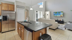 A modern kitchen with a stainless steel refrigerator, a built-in dishwasher, and a large island with a sink. There is a dining area with a gray sofa and a flat-screen TV mounted on the wall. - Villa Paradiso - South-facing Pool + Luxe Resort! (Kissimmee)