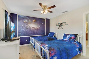 Un dormitorio con cama, escritorio con computadora, ventilador de techo, un mural en la pared y un pequeño modelo de avión.